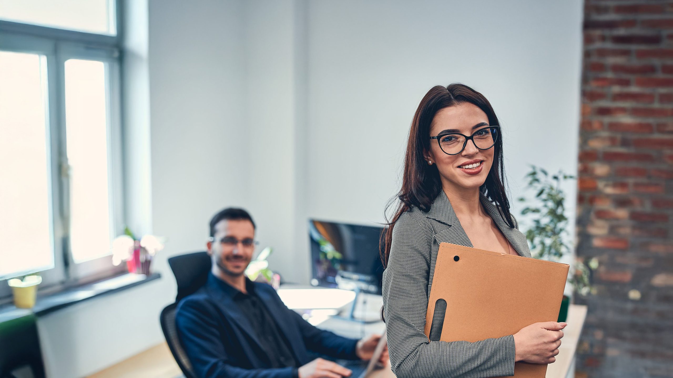 Back office employee smiling at the camera, wearing glasses, with a male colleague sitting in the background.